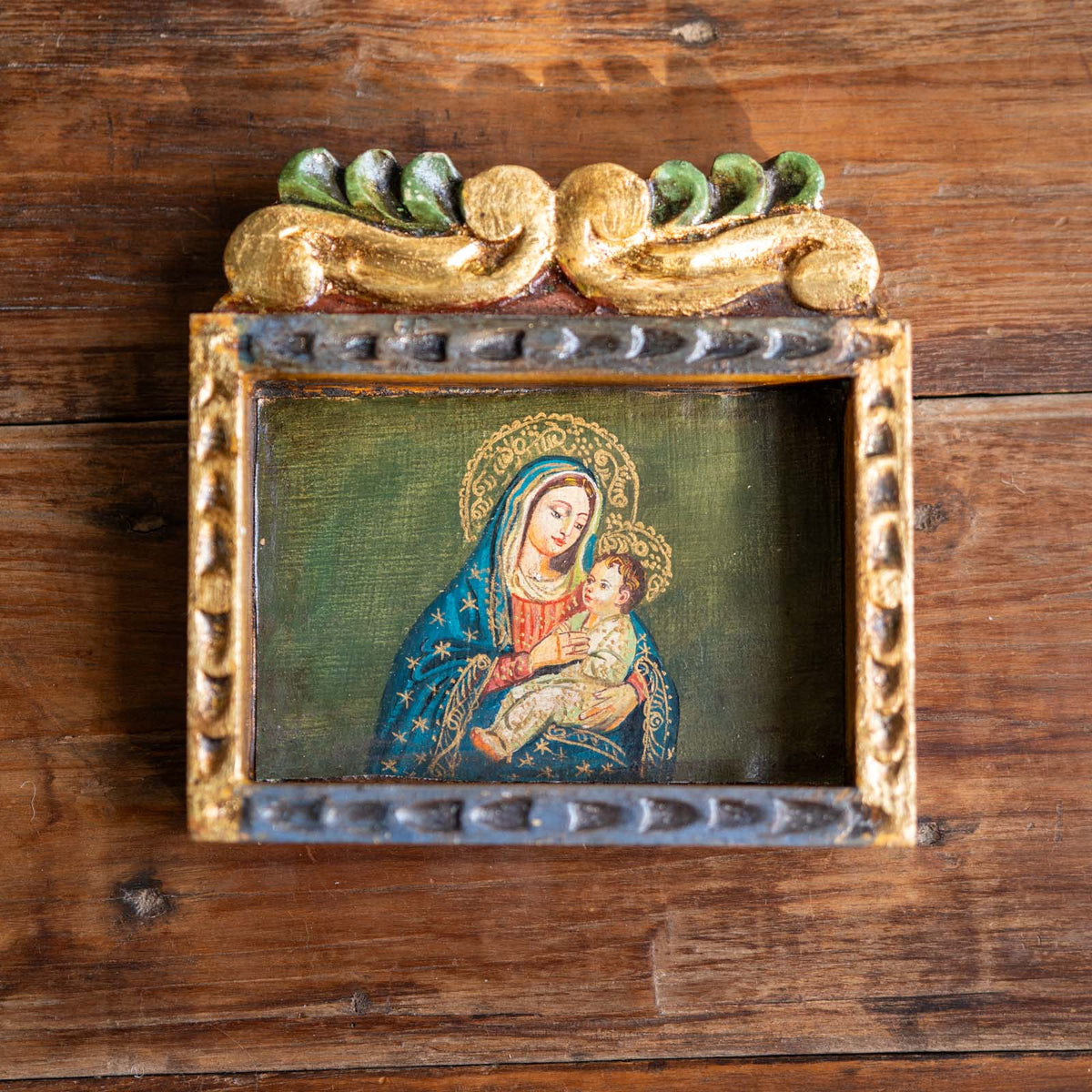 A small framed religious painting of the Virgin Mary holding the baby Jesus, mounted on a rustic dark wood background. The frame is ornate with a gold top scrollwork, a dark gray patterned lower border, and a blue robe with gold stars in the painting.