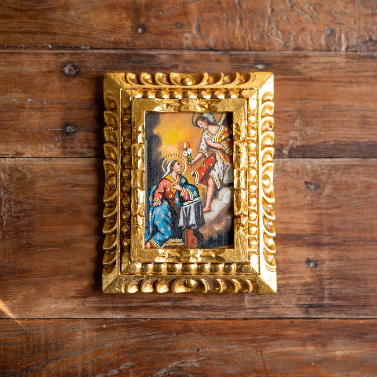 A small ornate gold-framed religious painting depicting the Annunciation of the Blessed Virgin Mary displayed on a rustic dark brown wooden background.