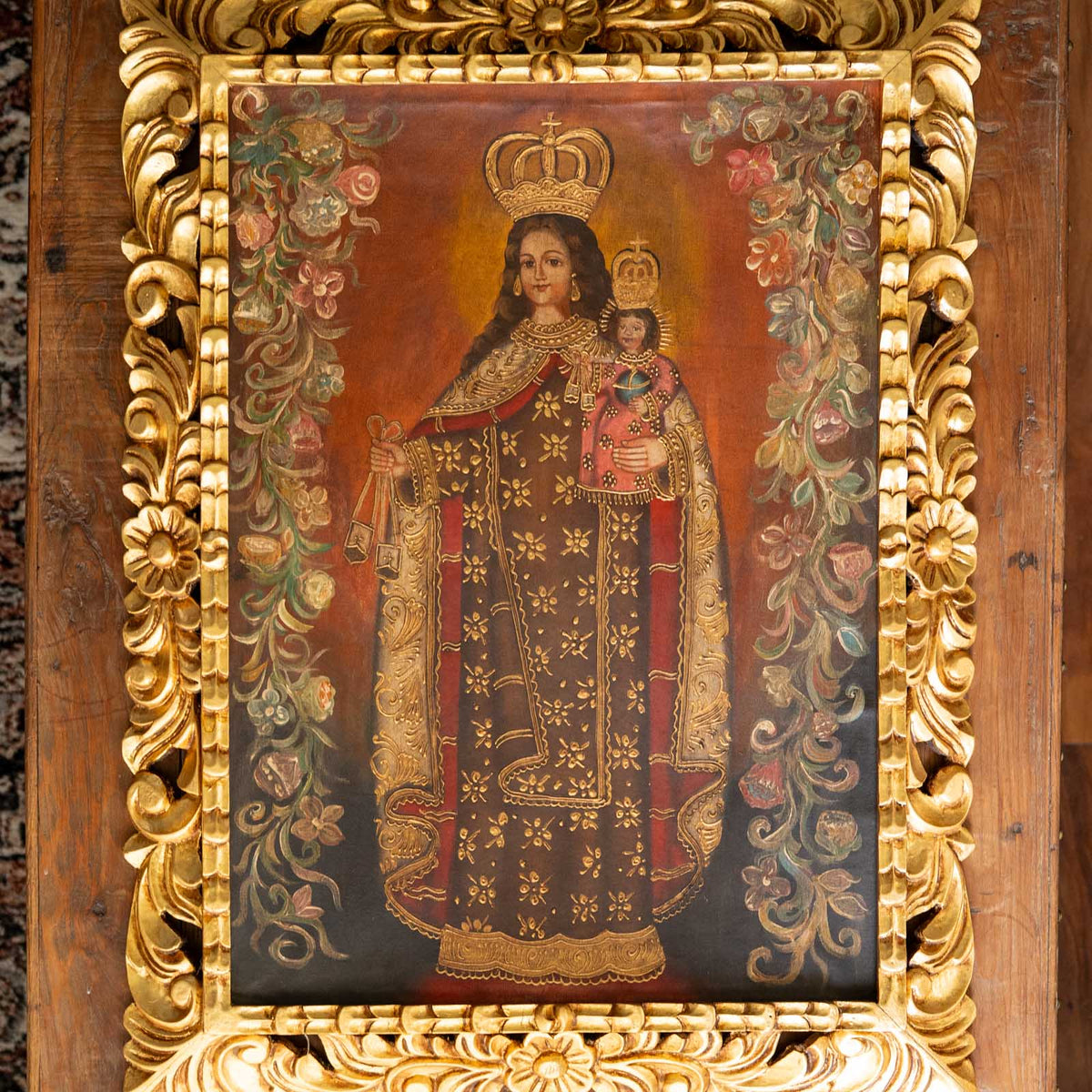 An ornate religious painting of the Virgin Mary holding the infant Jesus, enclosed in a highly decorative carved gold frame. The painting features Mary in a dark robe with gold floral patterns and red accents, and the infant Jesus wearing a crown.