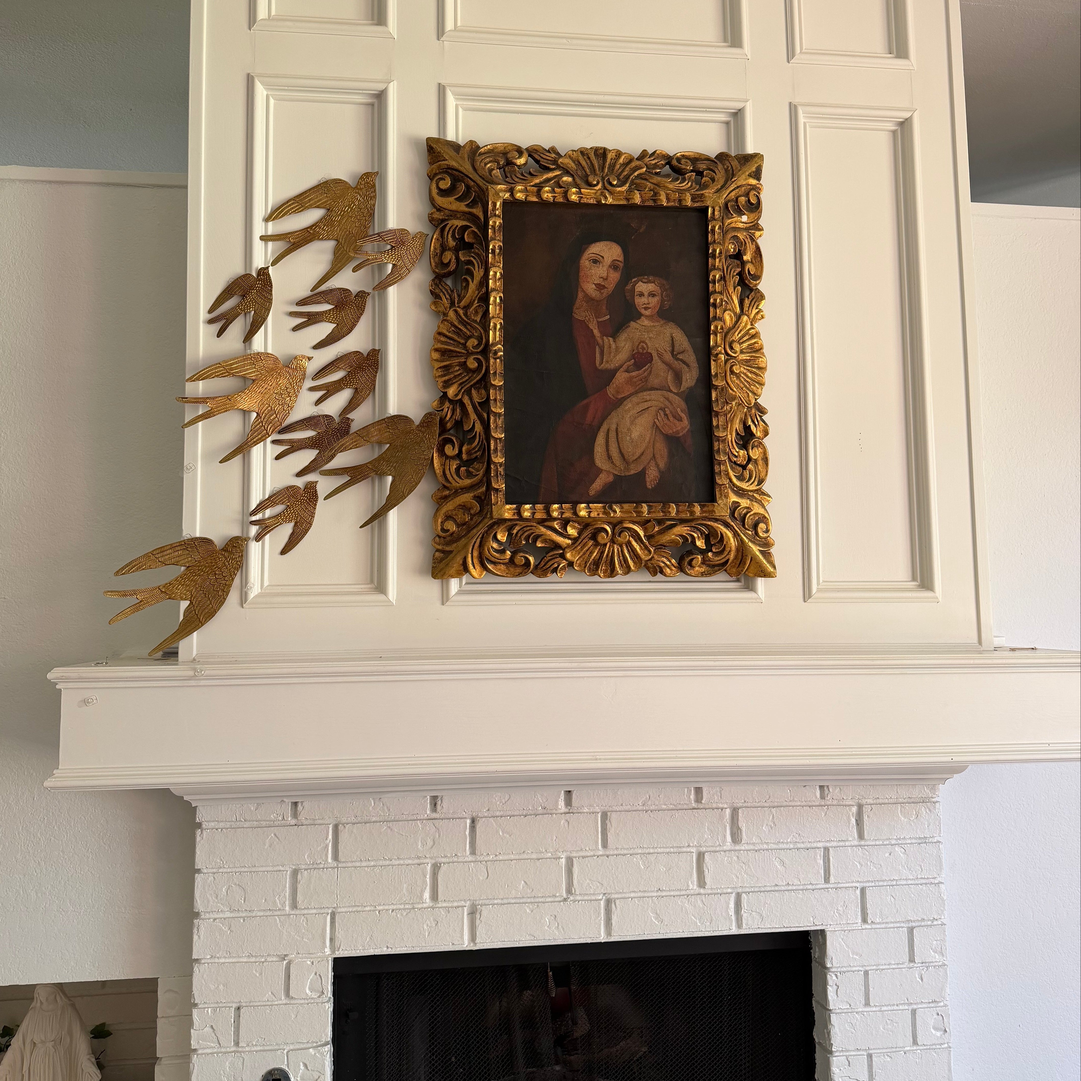 A white brick fireplace with a white mantel featuring a gold-framed religious painting and a gold bird sculpture on the left.