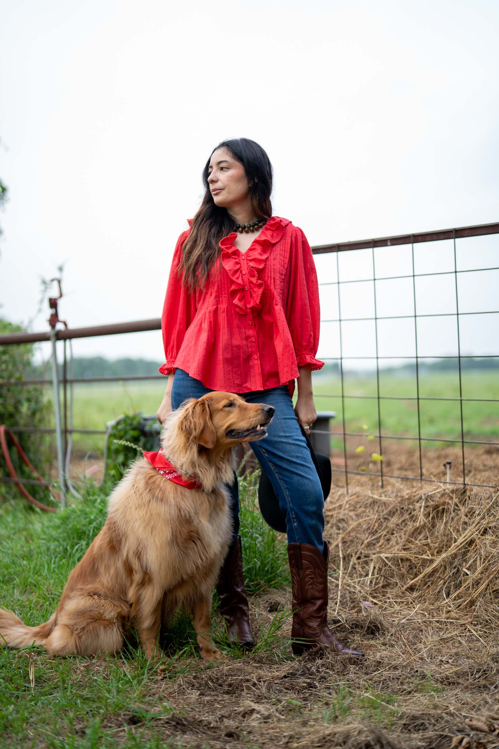 Woman in a red Rosa Blouse and blue jeans standing next to a golden retriever in a field.