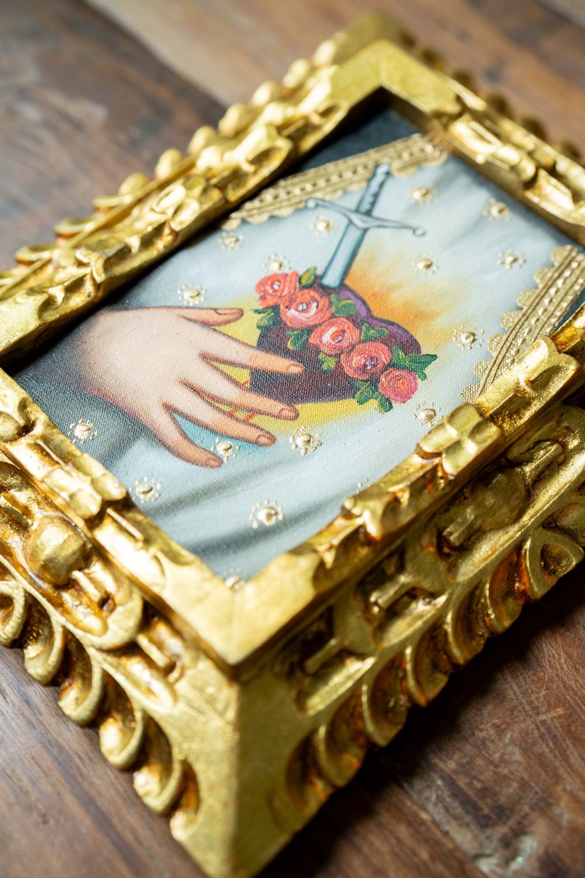 Decorative box with gold frame featuring a hand holding flowers and a cross on a wooden surface.
