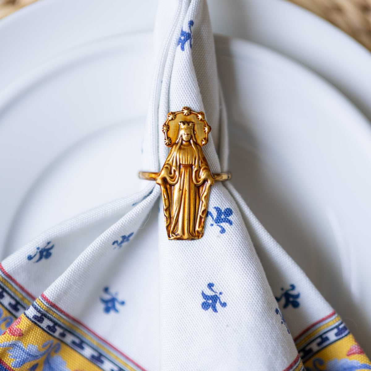 A gold-toned metal napkin ring shaped like the Virgin Mary holds a white patterned napkin with blue floral motifs and a multicolor border, arranged on a white plate.