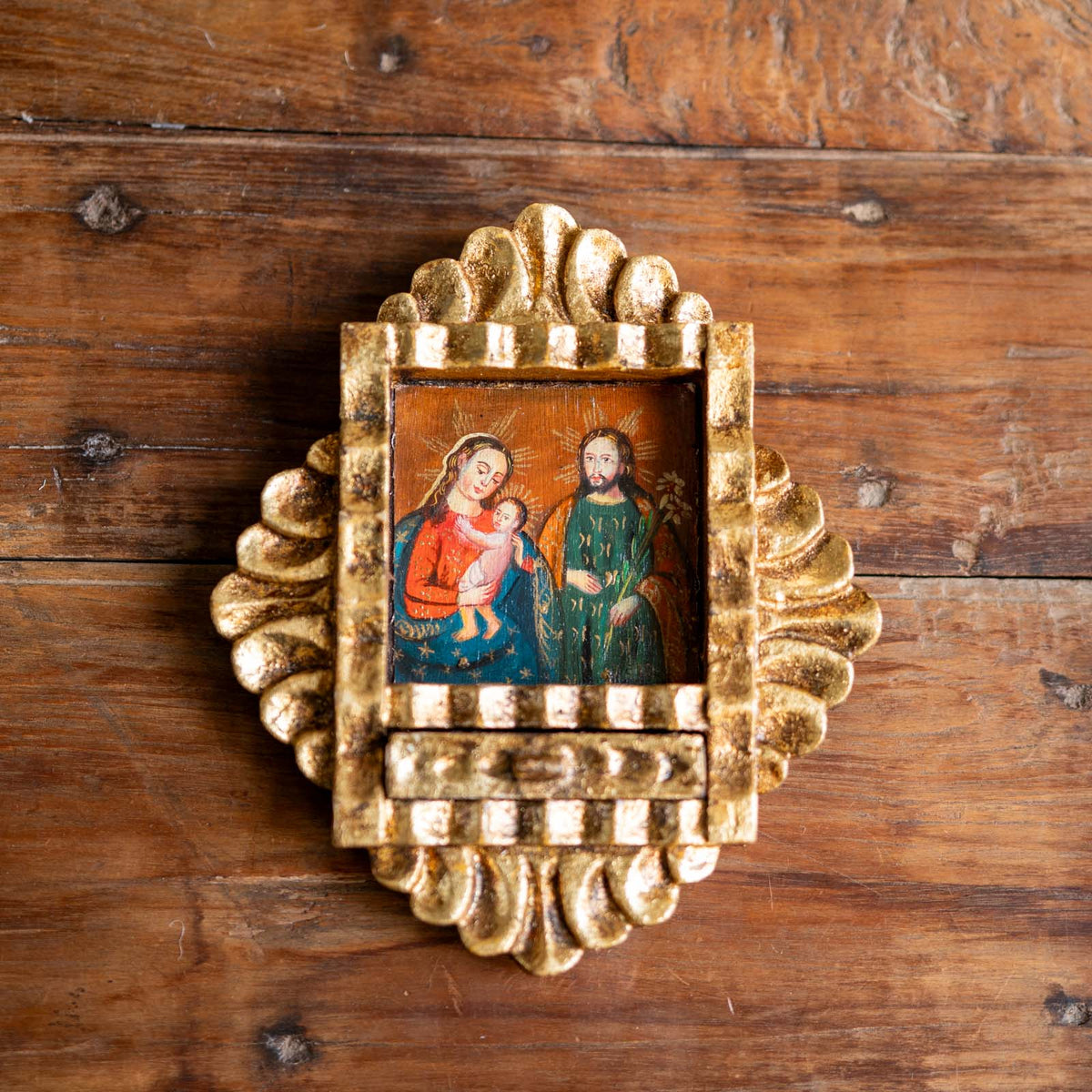 A small framed religious icon depicting the Holy Family, set in an ornate gold-toned frame with a textured, scalloped border, displayed on a rustic wooden surface.