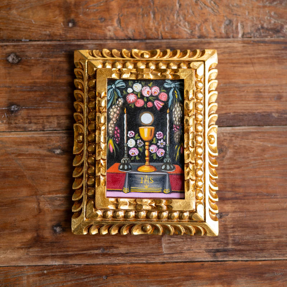 A small framed religious artwork depicting the Eucharist with a chalice, flowers, candles, and an open Bible labeled 'IHS', set in an ornate gold frame on a rustic wooden surface.