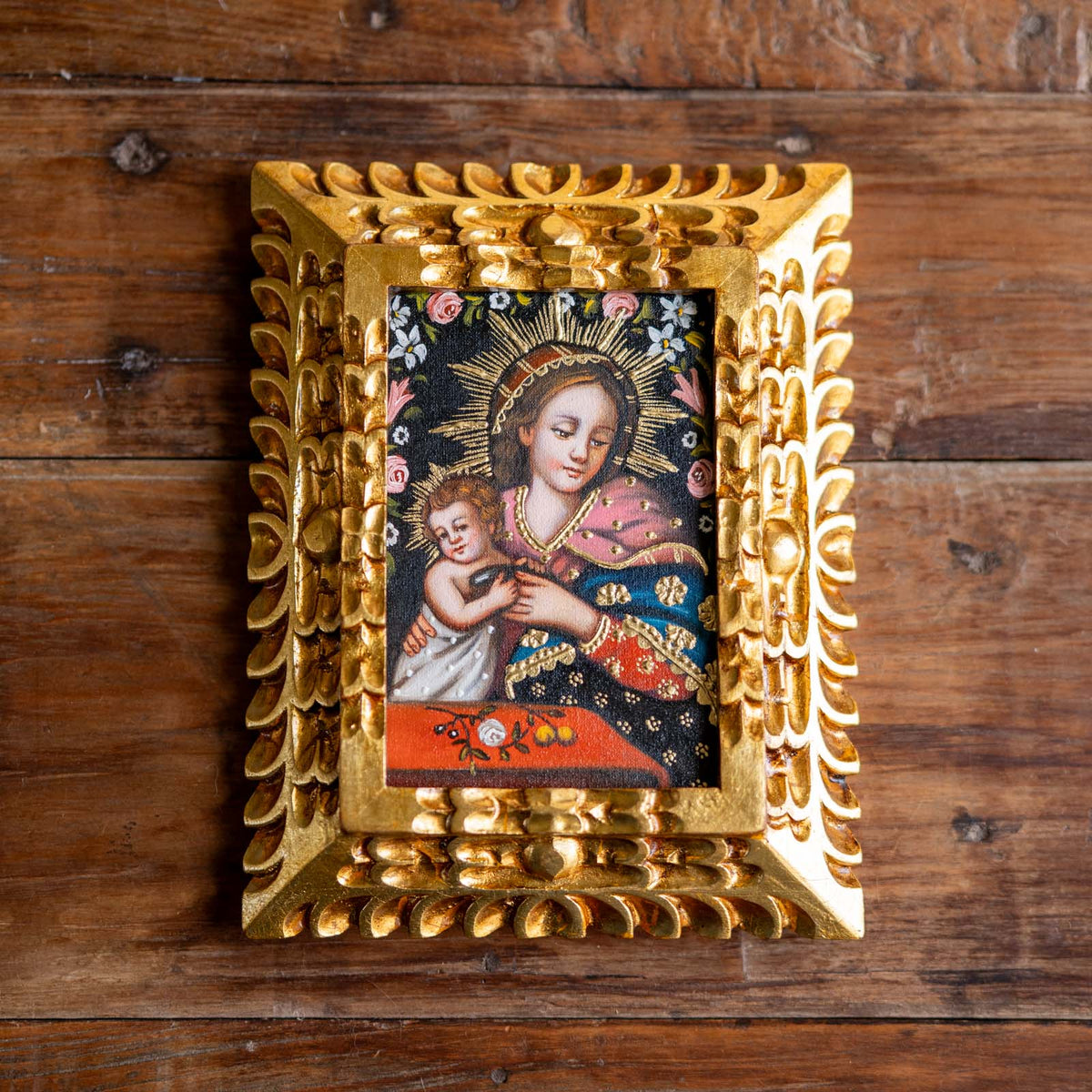 An ornate gold-colored frame holding a religious painting of the Virgin Mary holding the Christ child, displayed on a rustic dark wood background.