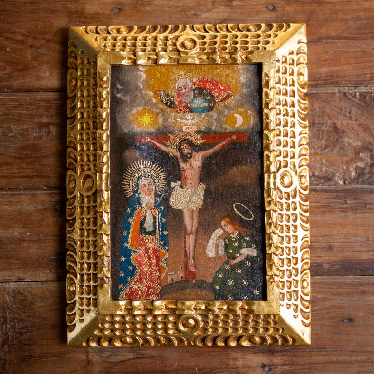 A framed religious painting depicting the Holy Trinity, featuring Jesus Christ, the Virgin Mary, and God, enclosed in an ornate, textured gold frame and displayed on a rustic wooden background.