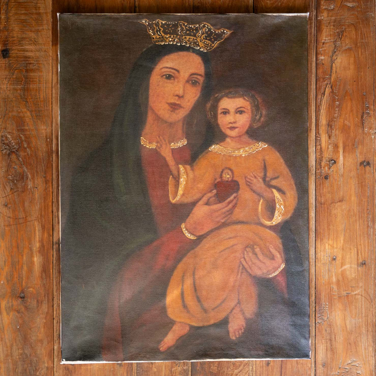 A rustic religious painting of the Virgin Mary holding the infant Jesus, mounted on a wooden board against a wooden wall. The painting features Mary in a dark green cloak and red dress with gold details, and Jesus in a yellow tunic, with a gold crown on Mary's head.