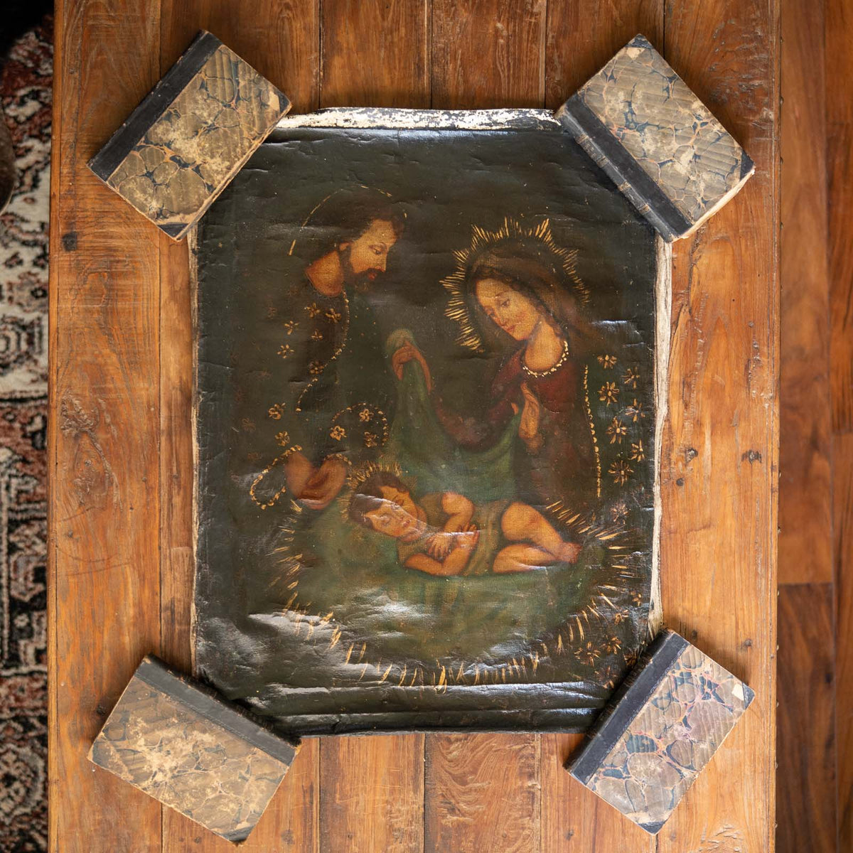 An antique religious painting of the Holy Family, mounted on a rustic wooden board with four decorative stone corner brackets, photographed from directly above on a wooden floor.