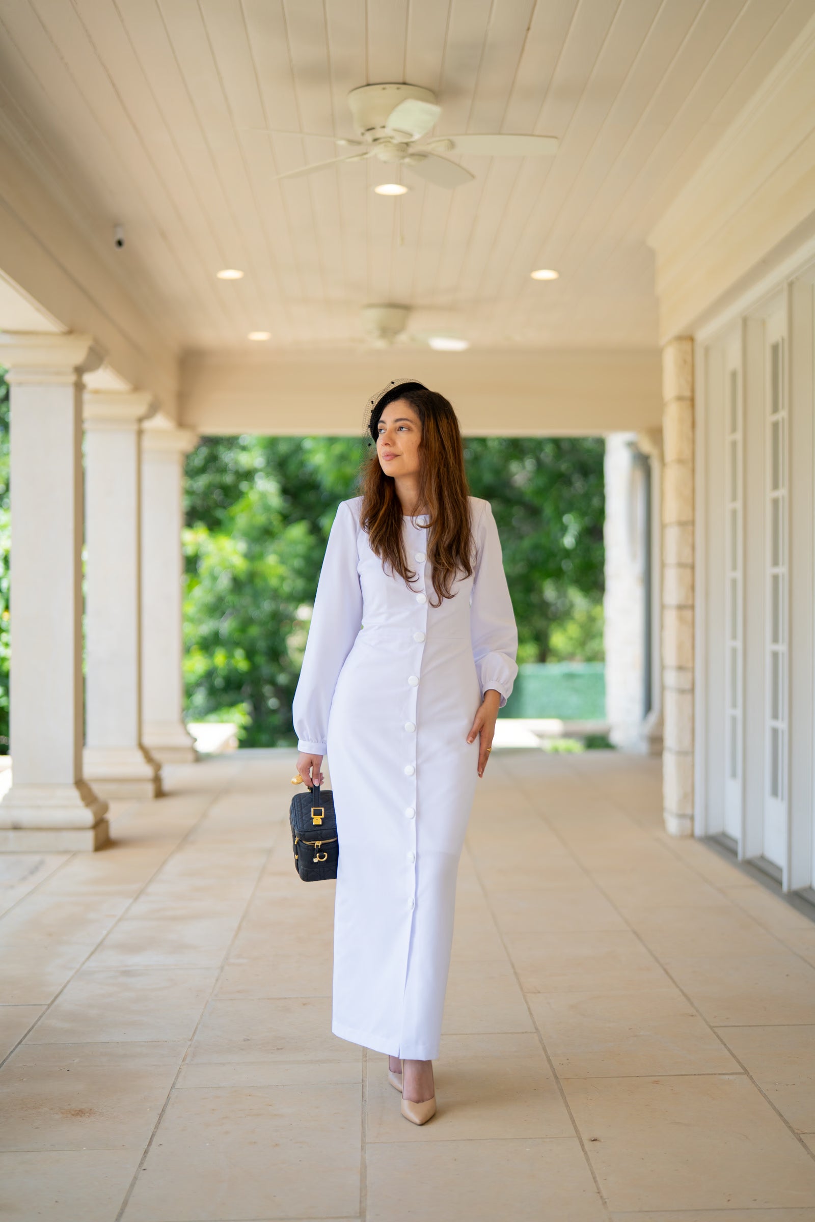 Woman in a white outfit standing in a covered outdoor area with columns and greenery.